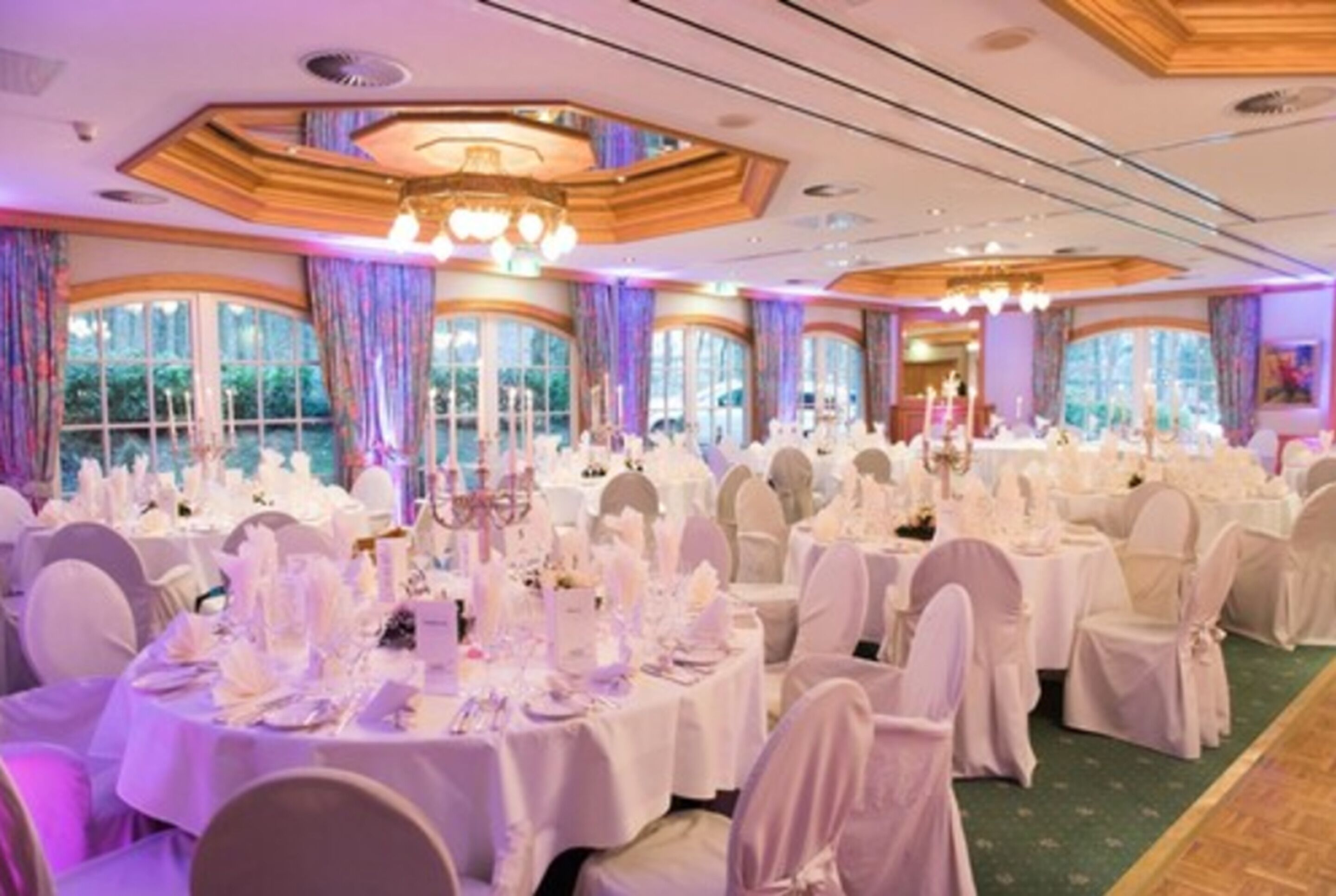 A banquet hall with round tables set for a formal event, large windows with curtains, and a chandelier.