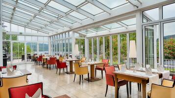 Un vaste espace intérieur aménagé pour les repas, avec de hauts plafonds et de grandes fenêtres, comprenant des tables et des chaises disposées pour faciliter les repas.
