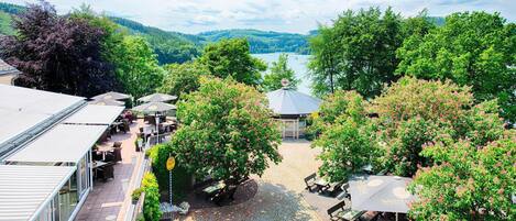 An outdoor seating area with tables and umbrellas, surrounded by lush greenery and a view of a lake and hills.