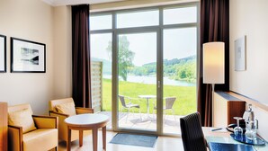 A hotel room with a large sliding glass door leading to a balcony, a small round table, two armchairs, a desk with a lamp, and a view of a green landscape. - Welcome Hotel Meschede/Hennesee (Meschede)