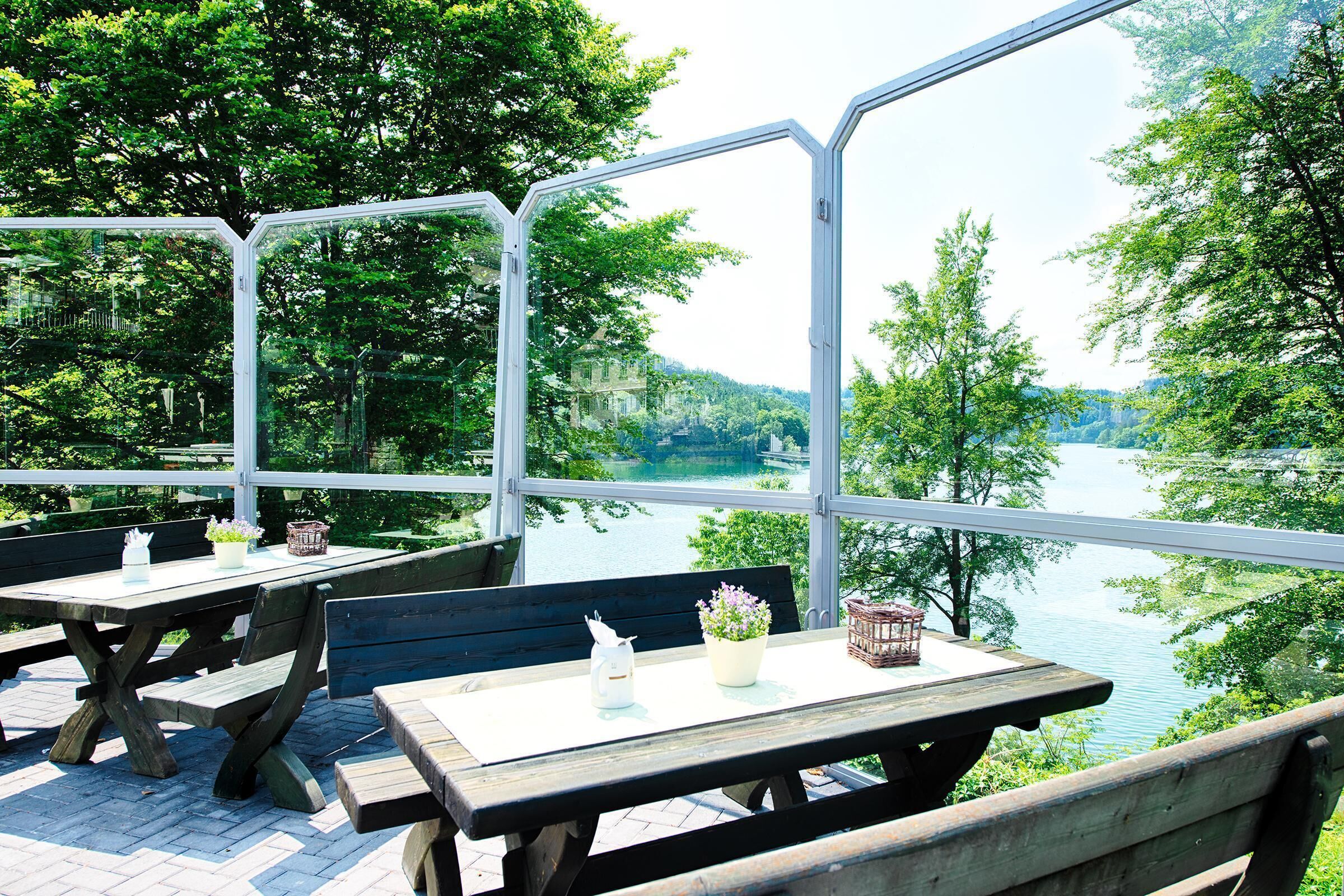 Une terrasse extérieure avec des tables et des bancs en bois, donnant sur un lac et entourée d’arbres.