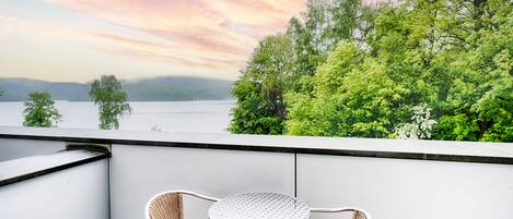 A balcony with wicker chairs and a small round table, overlooking a lake and lush greenery under a pink and blue sky.
