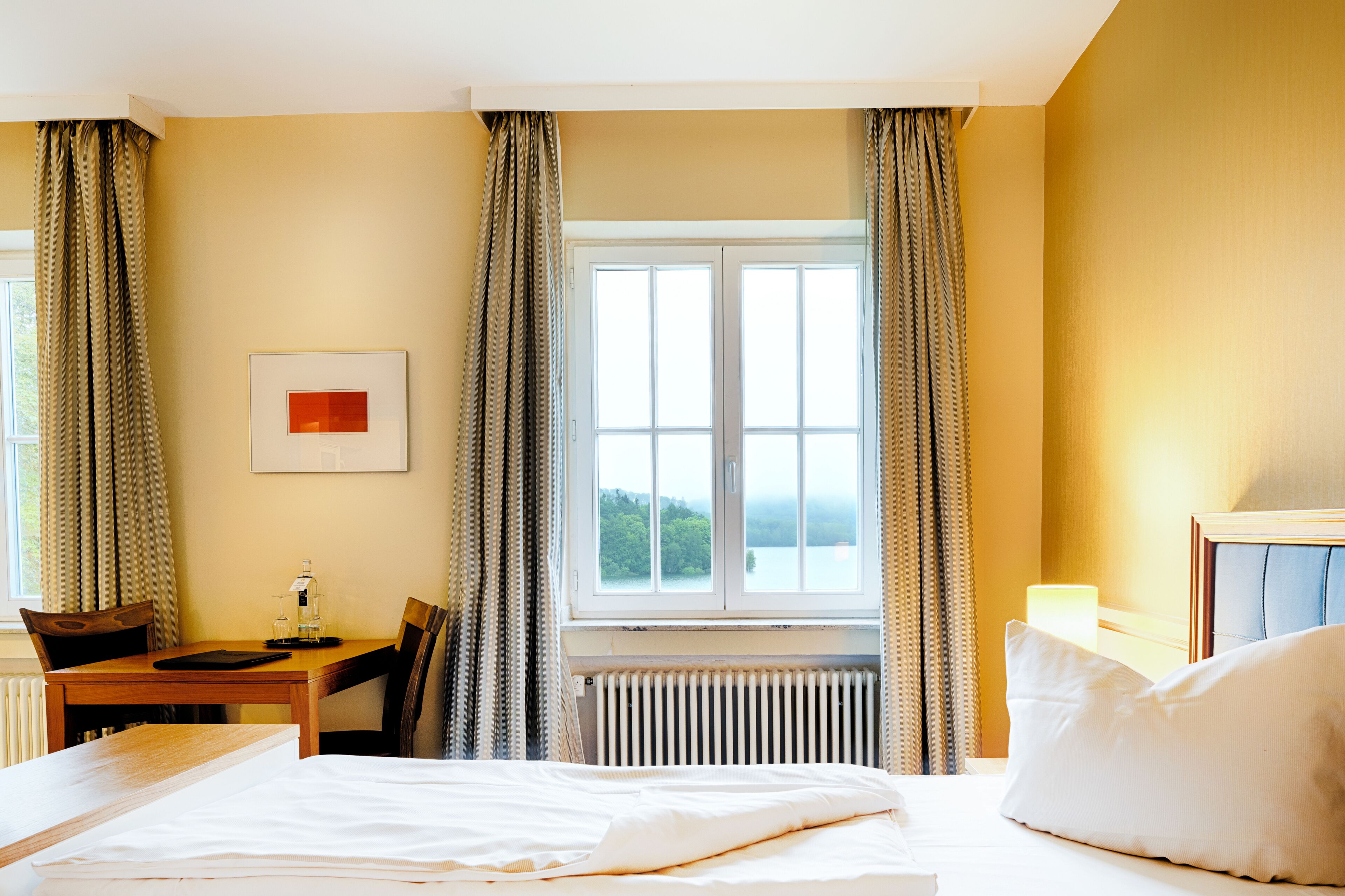 A hotel room with a bed, a desk with a chair, a window with curtains, and a radiator.