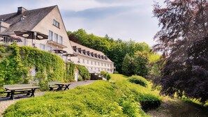 A large, multi-story building with a sloped roof, surrounded by lush greenery and trees. - Welcome Hotel Meschede/Hennesee (Meschede)