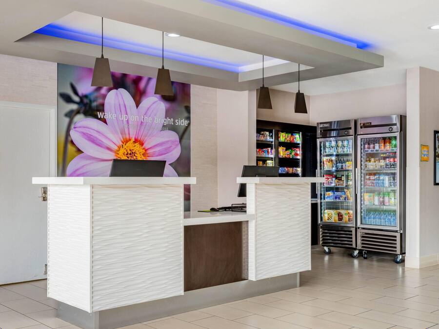 Un área de recepción moderna con un mostrador blanco, iluminación colgante, una pantalla digital con una imagen floral y un refrigerador de bebidas.
