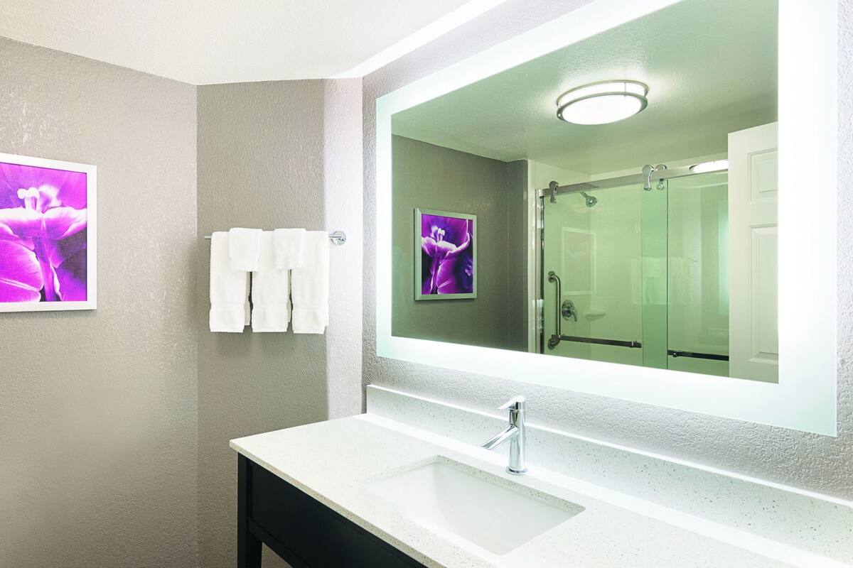 Une salle de bain avec un lavabo blanc, un grand miroir et une douche visible dans le reflet.
