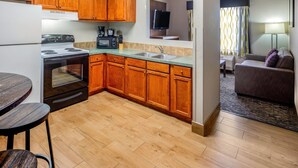 A compact kitchen with wooden cabinets, a stove, microwave, and a sink. There is a dining area with a table and chairs, and a living room with a sofa and a TV.