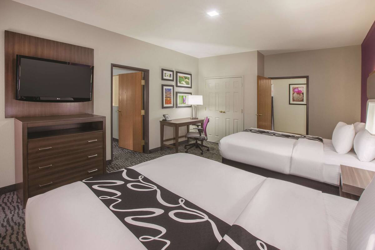 Une chambre d’hôtel comprenant un grand lit, un lit plus petit, un bureau avec une chaise, un téléviseur fixé sur un panneau en bois et une porte donnant sur une autre pièce.