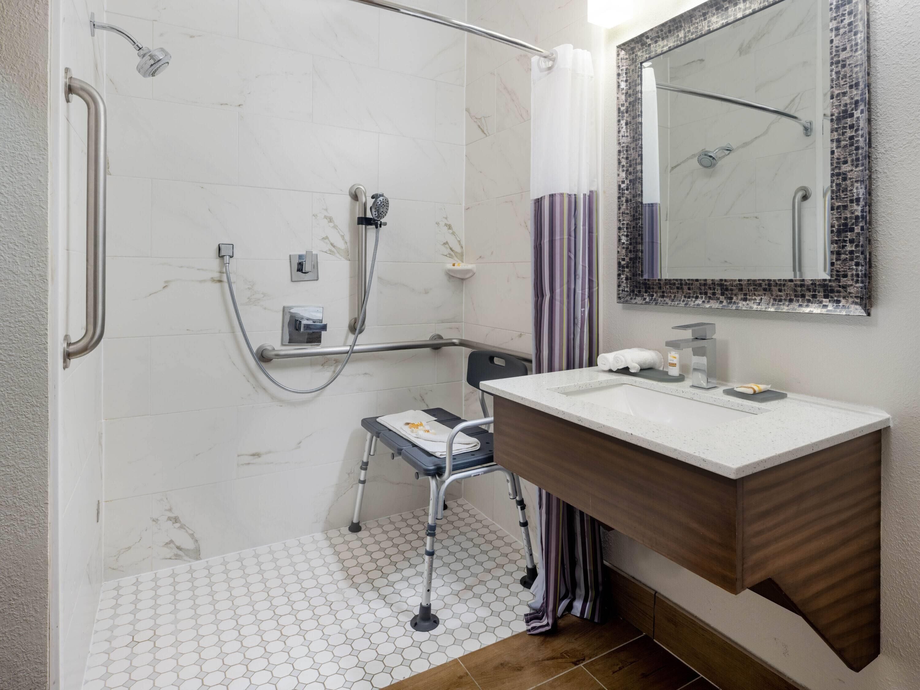 A bathroom with a white tiled shower, a wooden vanity, a mirror with a mosaic frame, and a wheelchair-accessible shower chair.