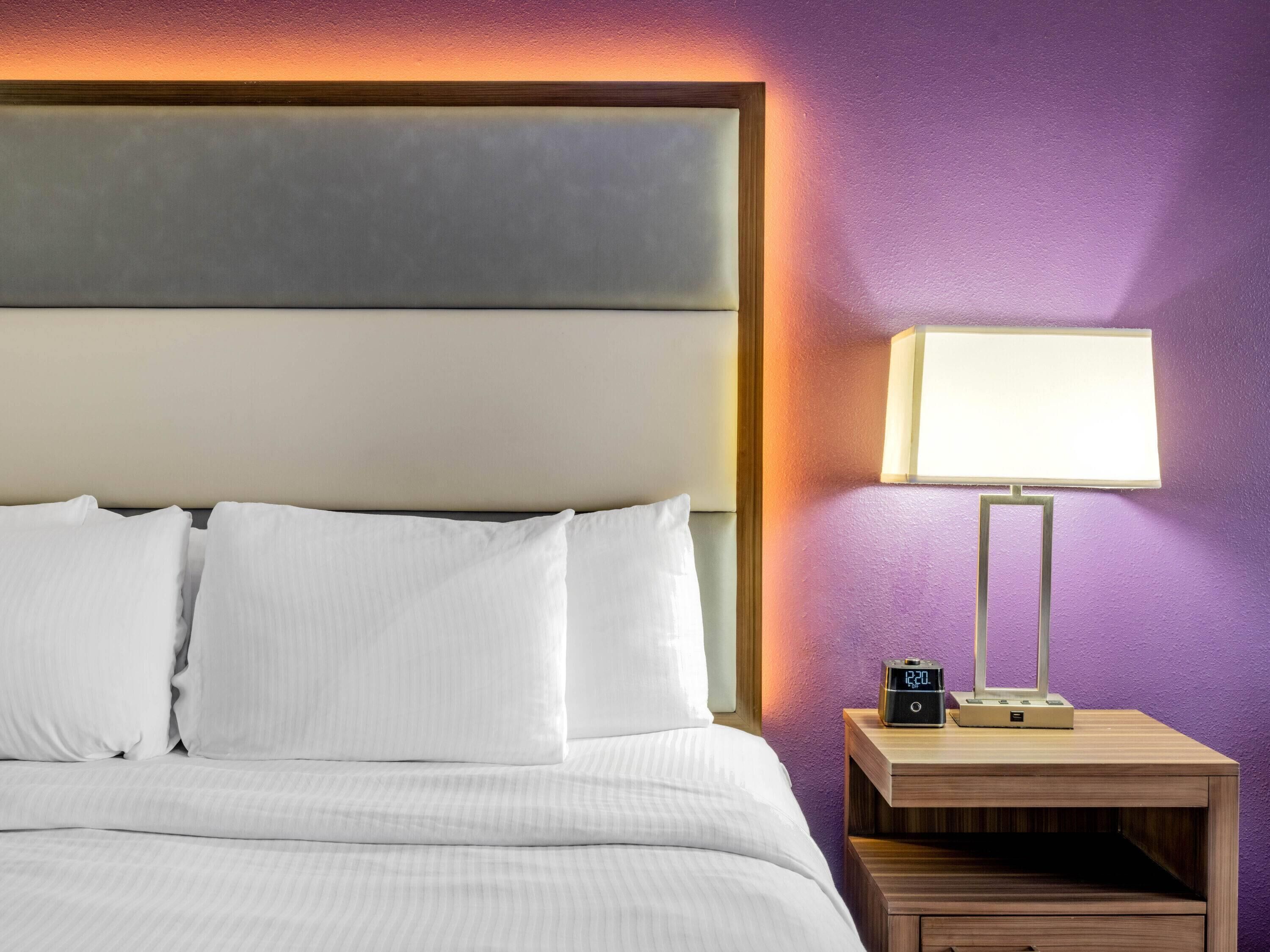 A neatly made bed with white linens and pillows, a bedside table with a lamp and a clock, and a purple wall.