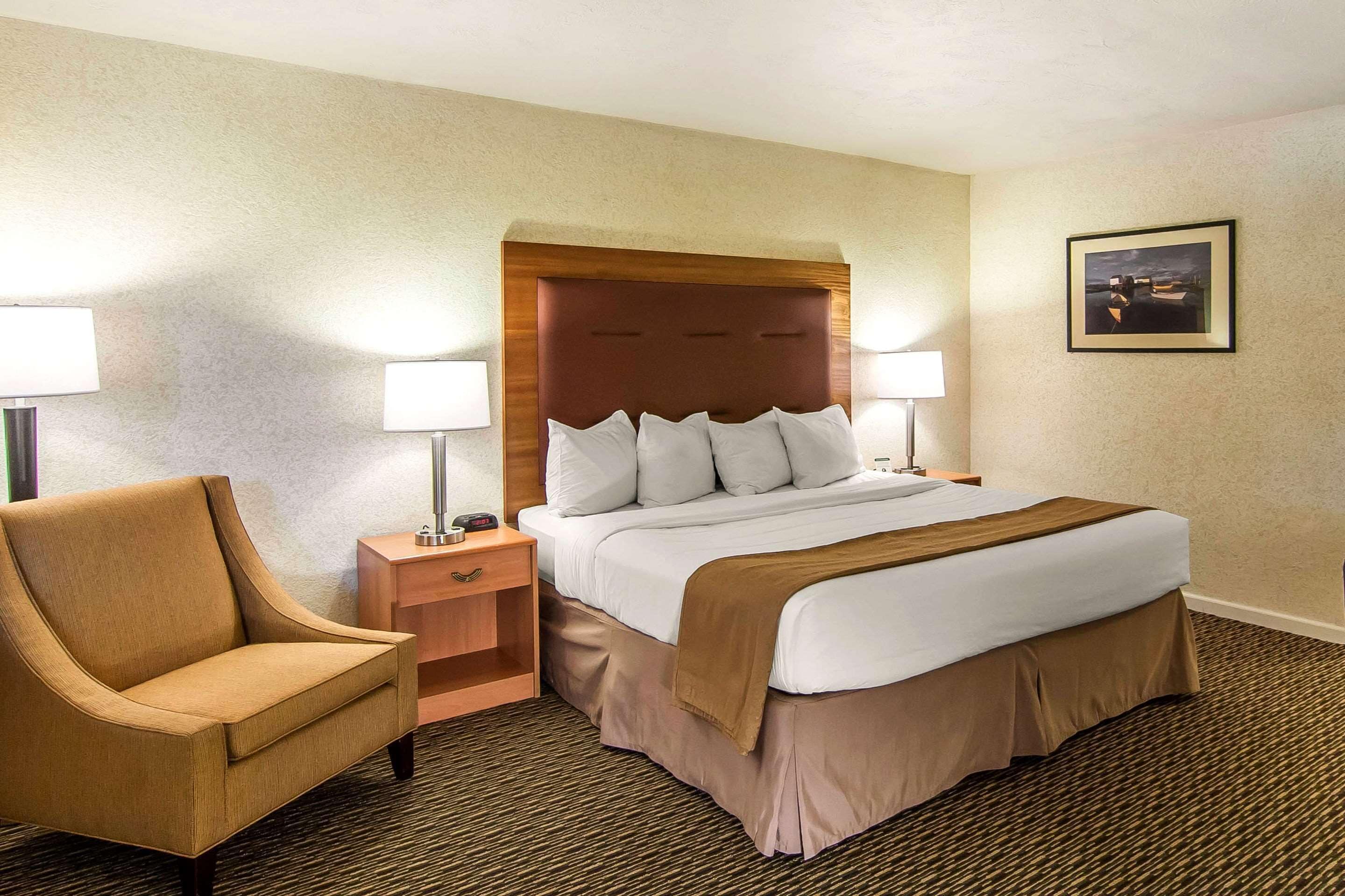 A hotel room with a large bed, two bedside lamps, a wooden nightstand, a brown armchair, and a framed picture on the wall.
