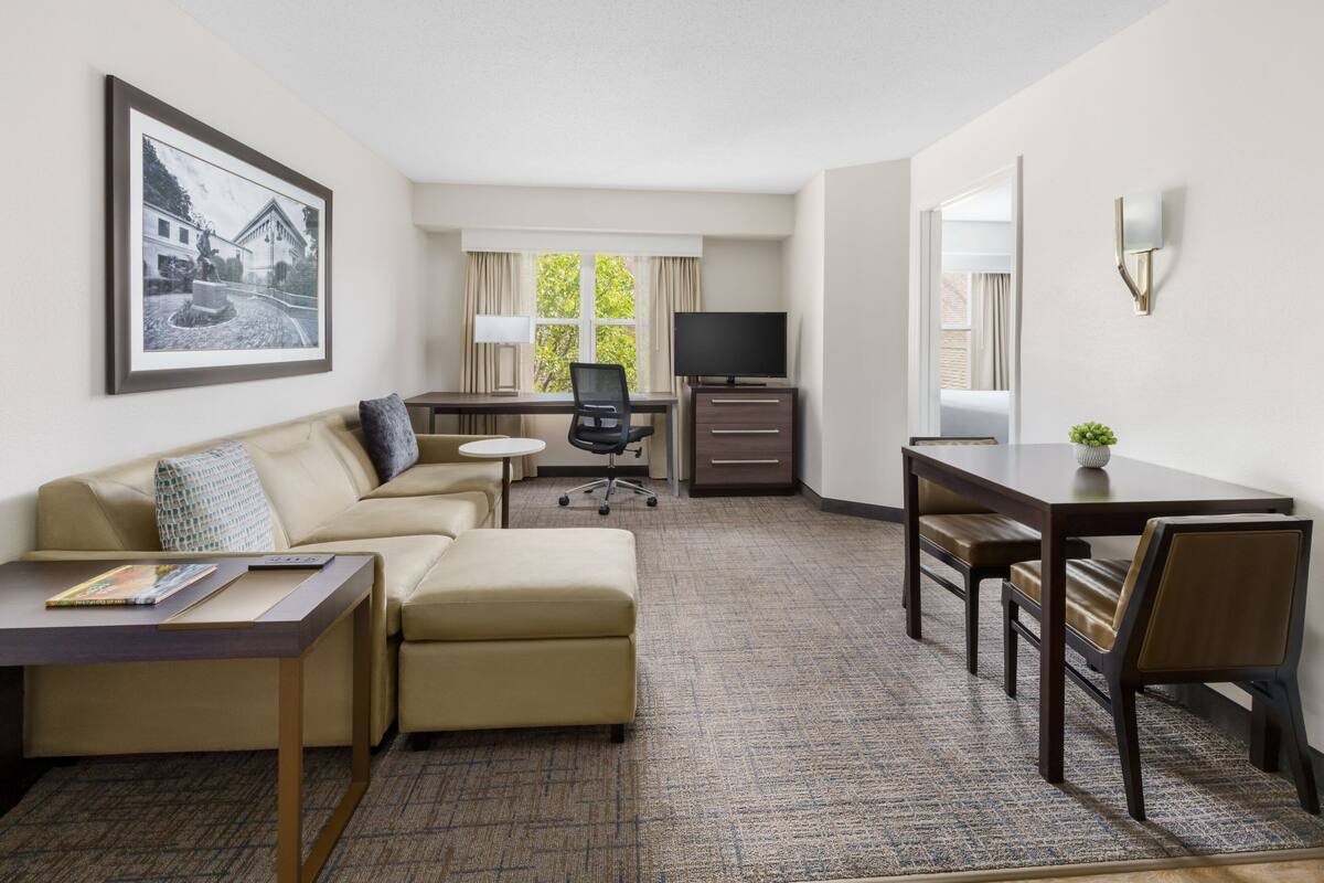 Ein Hotelzimmer mit einer Couch, einem kleinen Tisch, einem Schreibtisch mit Stuhl, einem Fernseher und einem gerahmten Bild an der Wand.