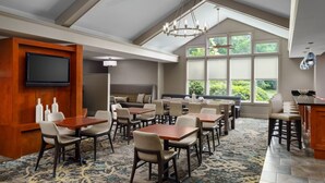 A dining area with multiple tables and chairs, a flat-screen TV mounted on a wooden panel, and large windows offering a view of greenery. - Residence Inn by Marriott Boston-Franklin (Franklin)