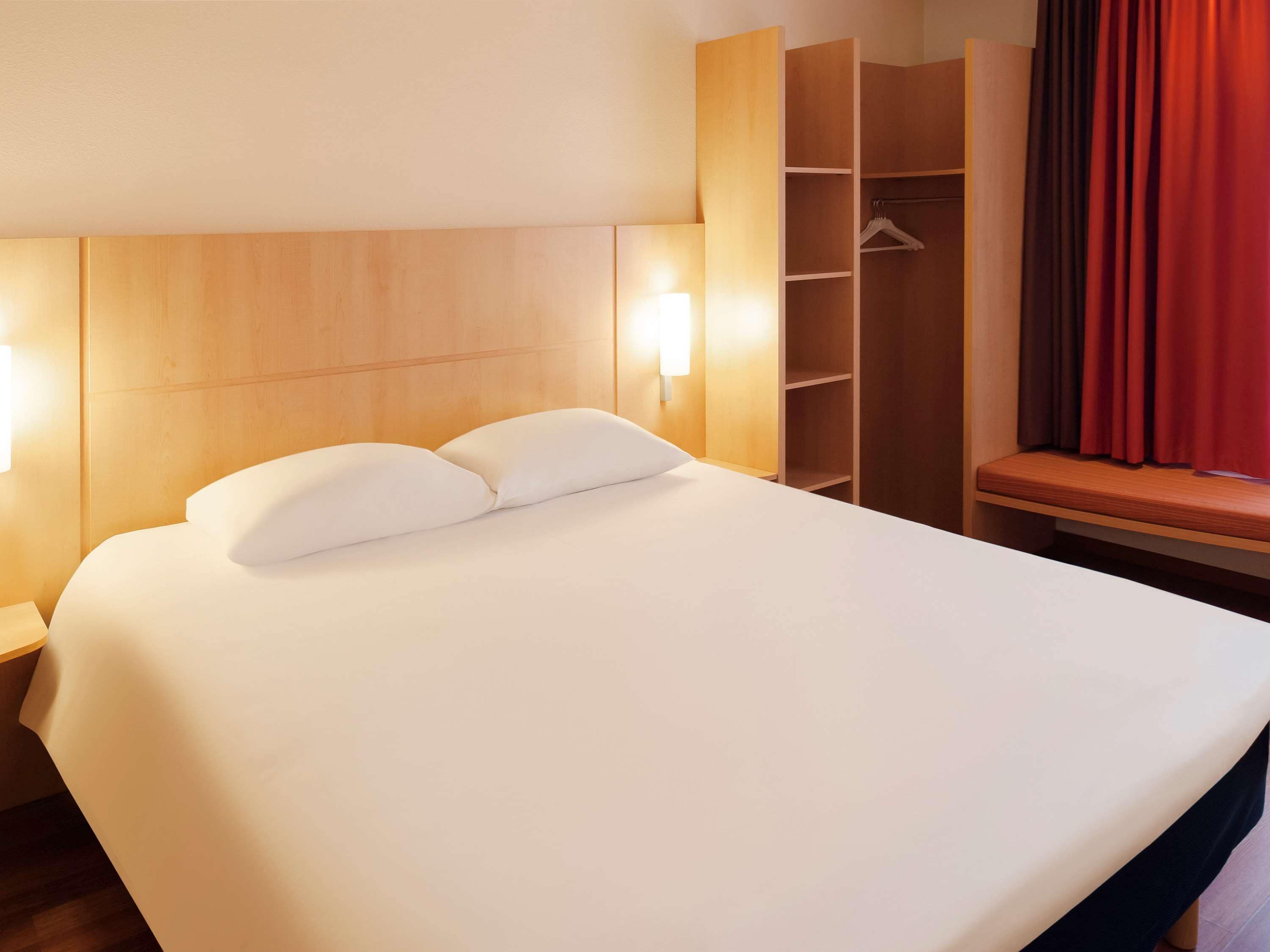 A hotel room with a large bed, a wooden wardrobe, a bench, and a window with red curtains.