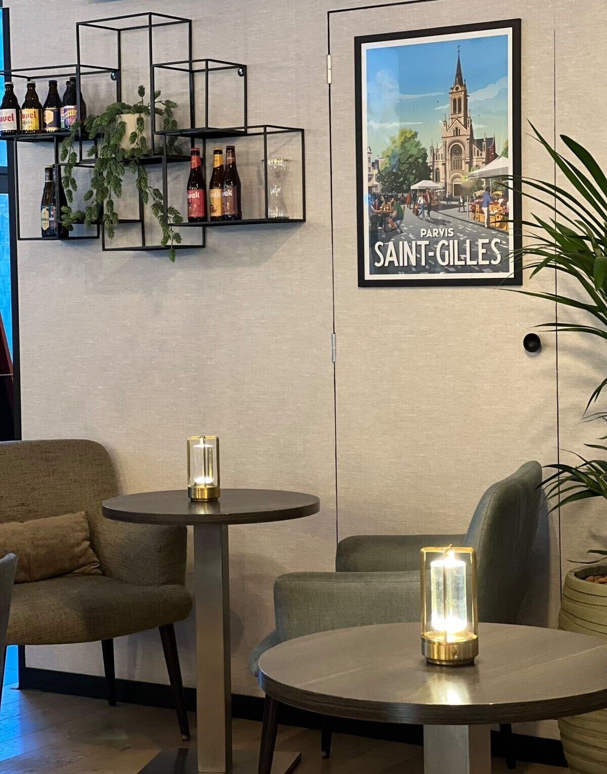 Un interior acogedor con dos mesas redondas, un cartel enmarcado de Saint-Gilles y una estantería con botellas y plantas.