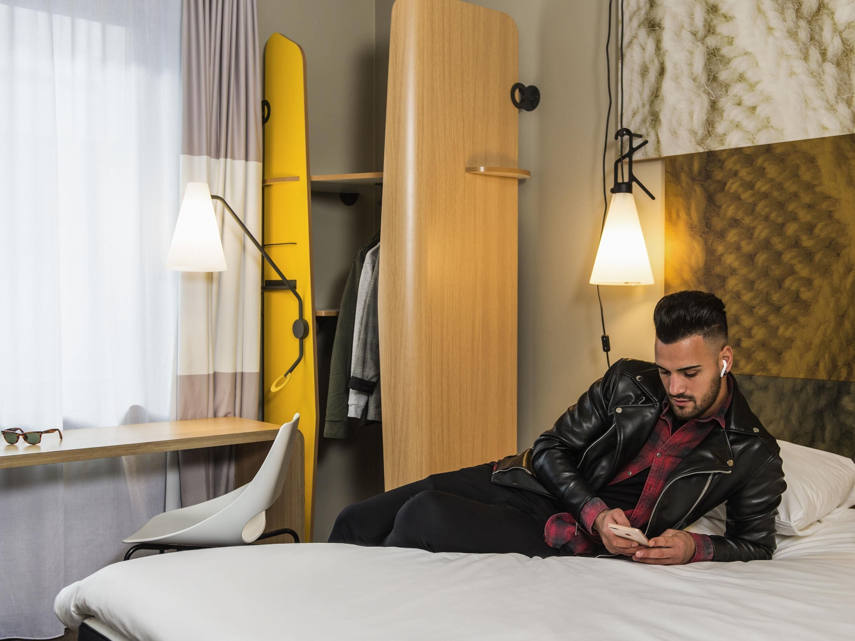 A hotel room with a bed, a desk, a chair, a surfboard, and a wardrobe.