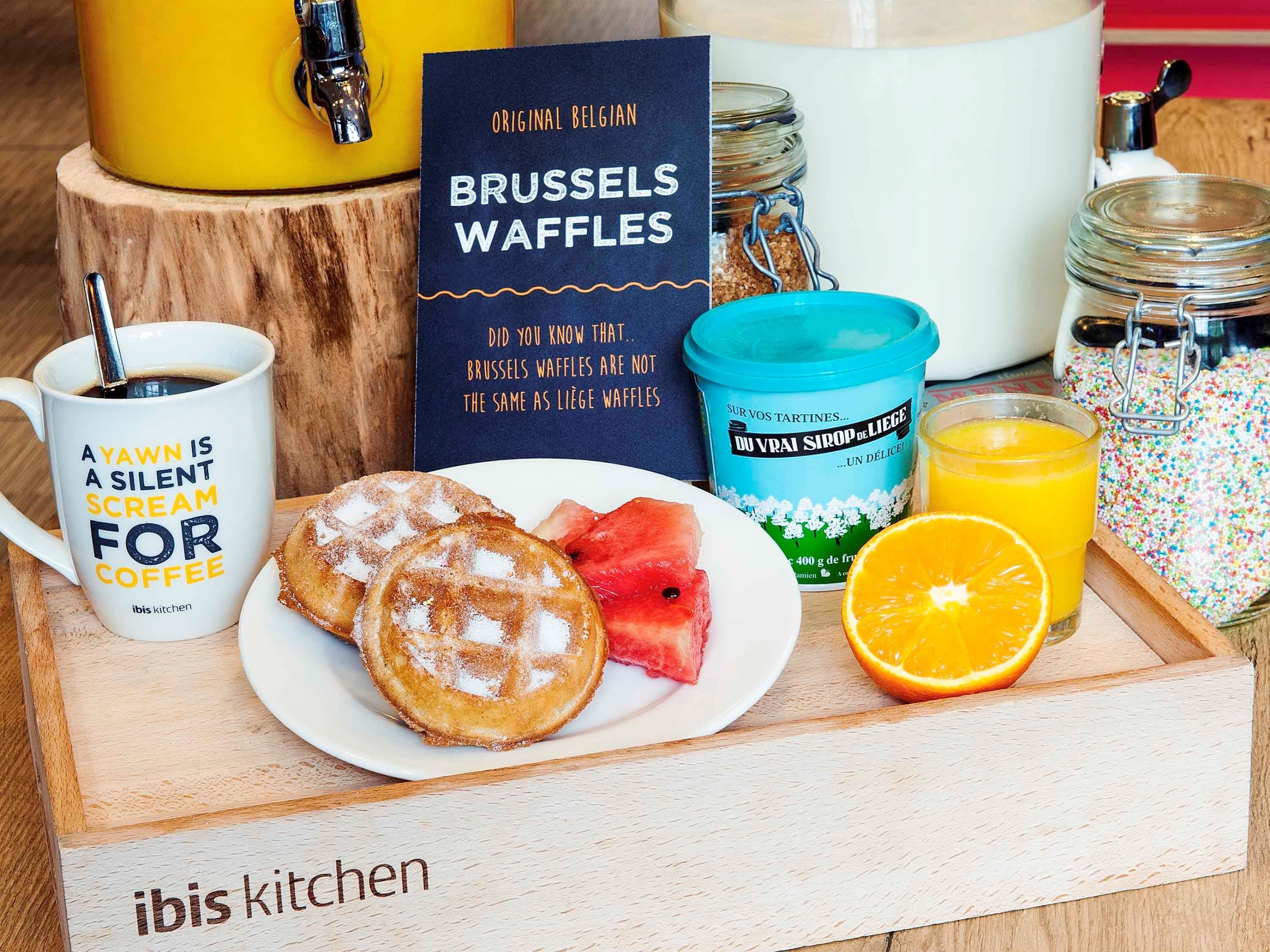 Bandeja de desayuno con waffles, una taza de café, tarjeta informativa sobre waffles de Bruselas, un tarro de jarabe y un vaso de jugo de naranja.