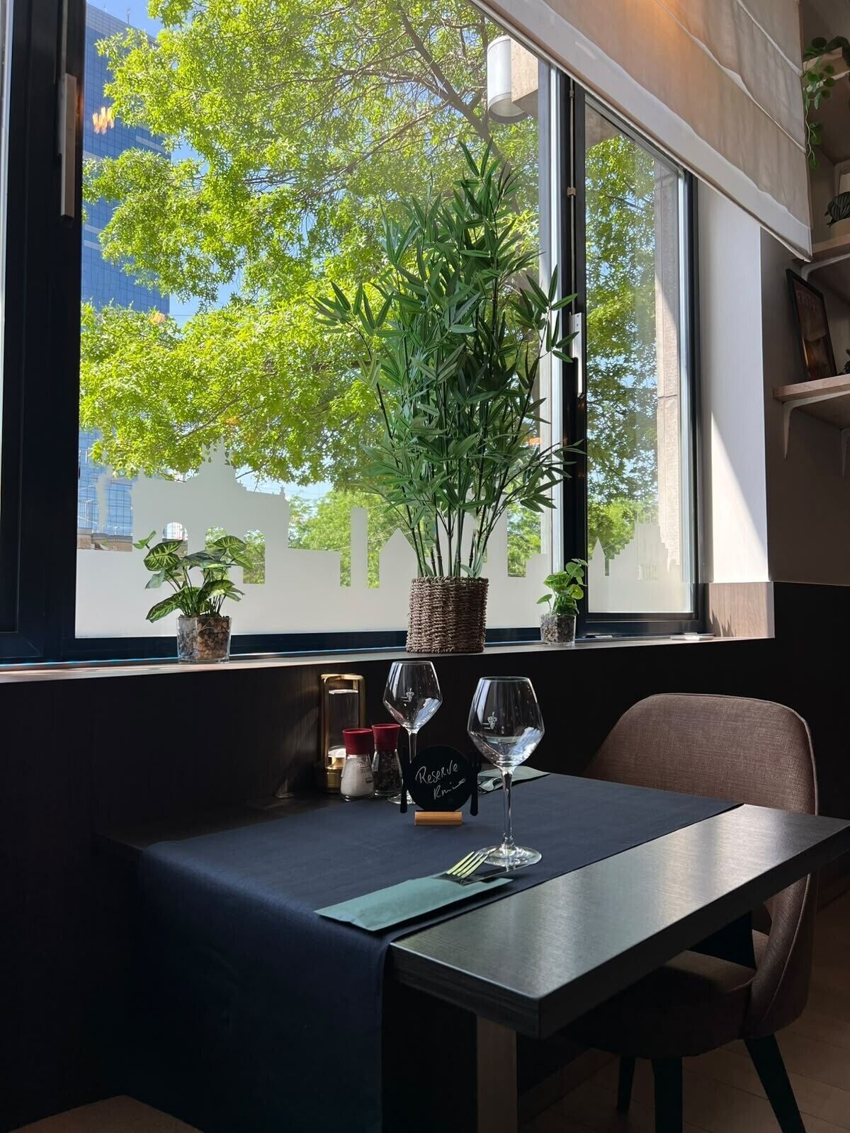 A table with a dark tablecloth, two wine glasses, a menu, and a small plant by a window with a view of trees and buildings.