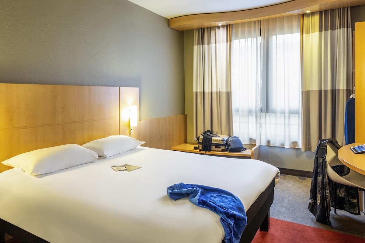 Una habitación de hotel con cama, escritorio, silla, lámpara y ventana con cortinas.