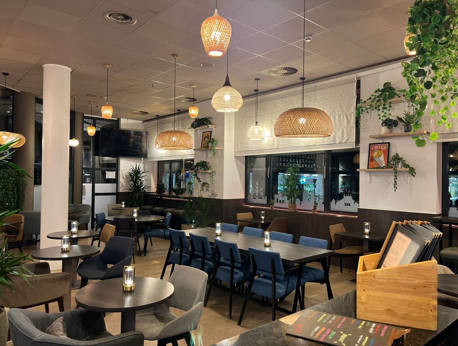 Interior de restaurante com mesas redondas, cadeiras azuis, luminárias de vime pendentes e plantas em vasos.