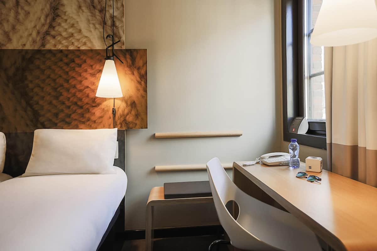 Habitación de hotel con una cama, un escritorio con lámpara, una silla, una ventana y una estantería en la pared.