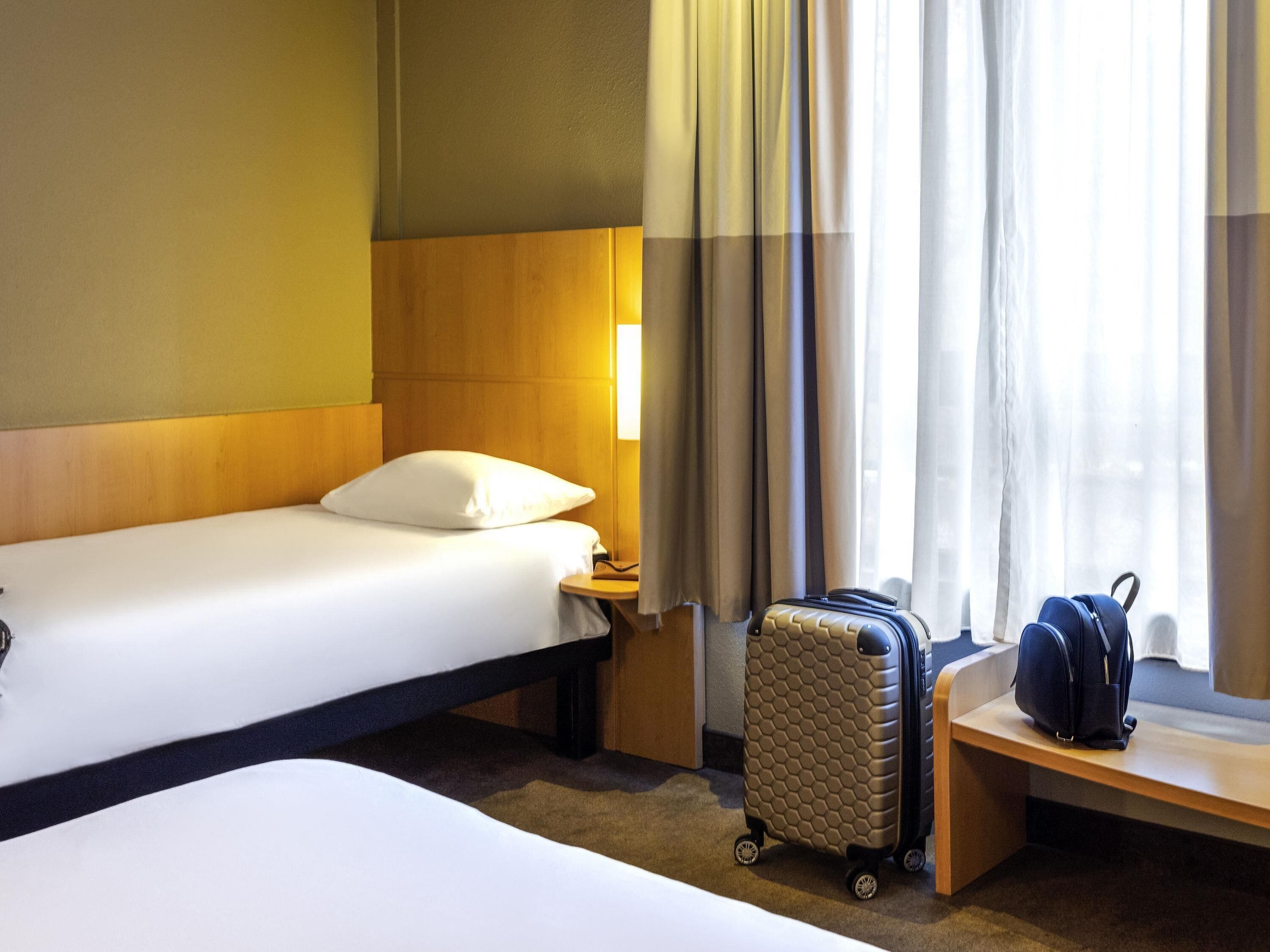 A hotel room with two beds, a suitcase, and a backpack on a bench by the window.