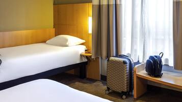 Une chambre d’hôtel avec deux lits, une valise et un sac à dos posés sur un banc près de la fenêtre.