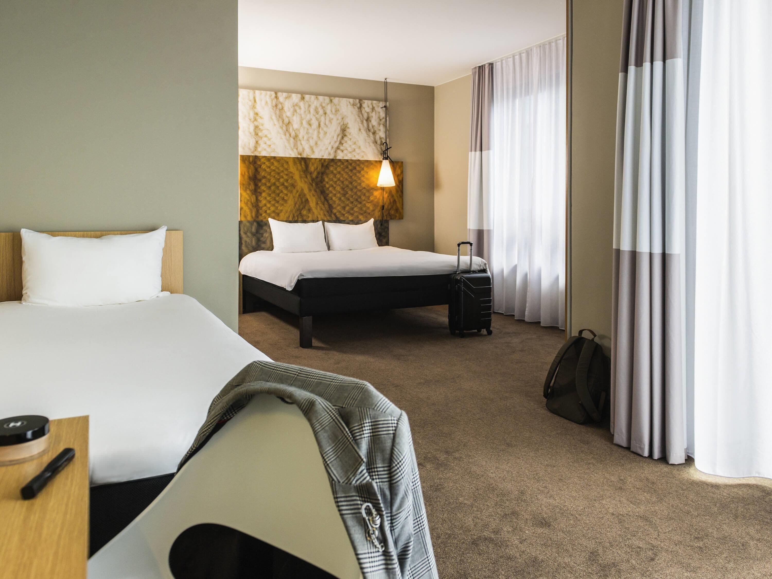 A hotel room with two beds, a headboard with a light fixture, a suitcase, and a desk with a remote control.