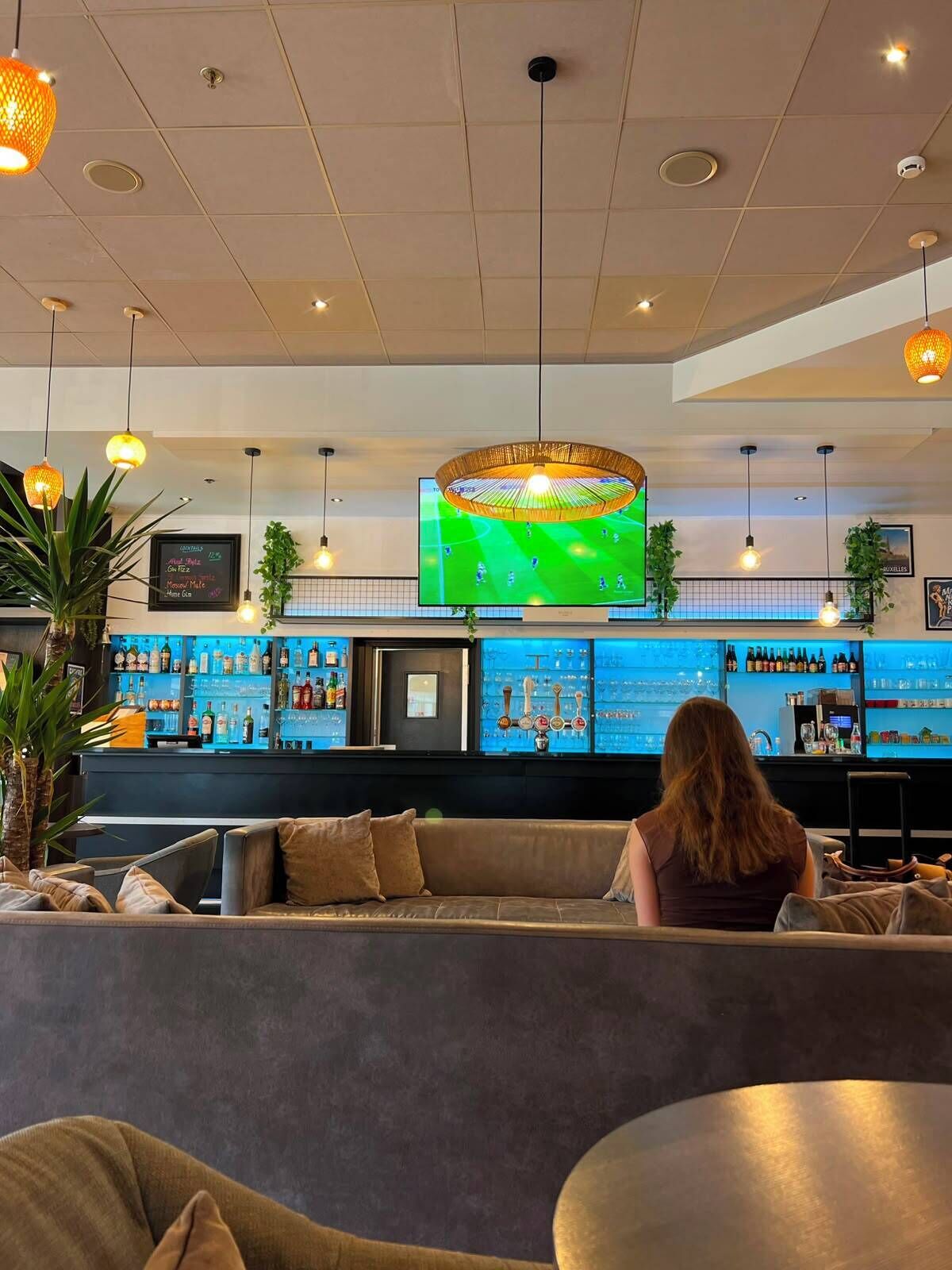 Eine Bar mit Fernsehbild eines Fußballspiels, einer Theke voller Flaschen und einer Sitzecke mit Sofa und Tisch.