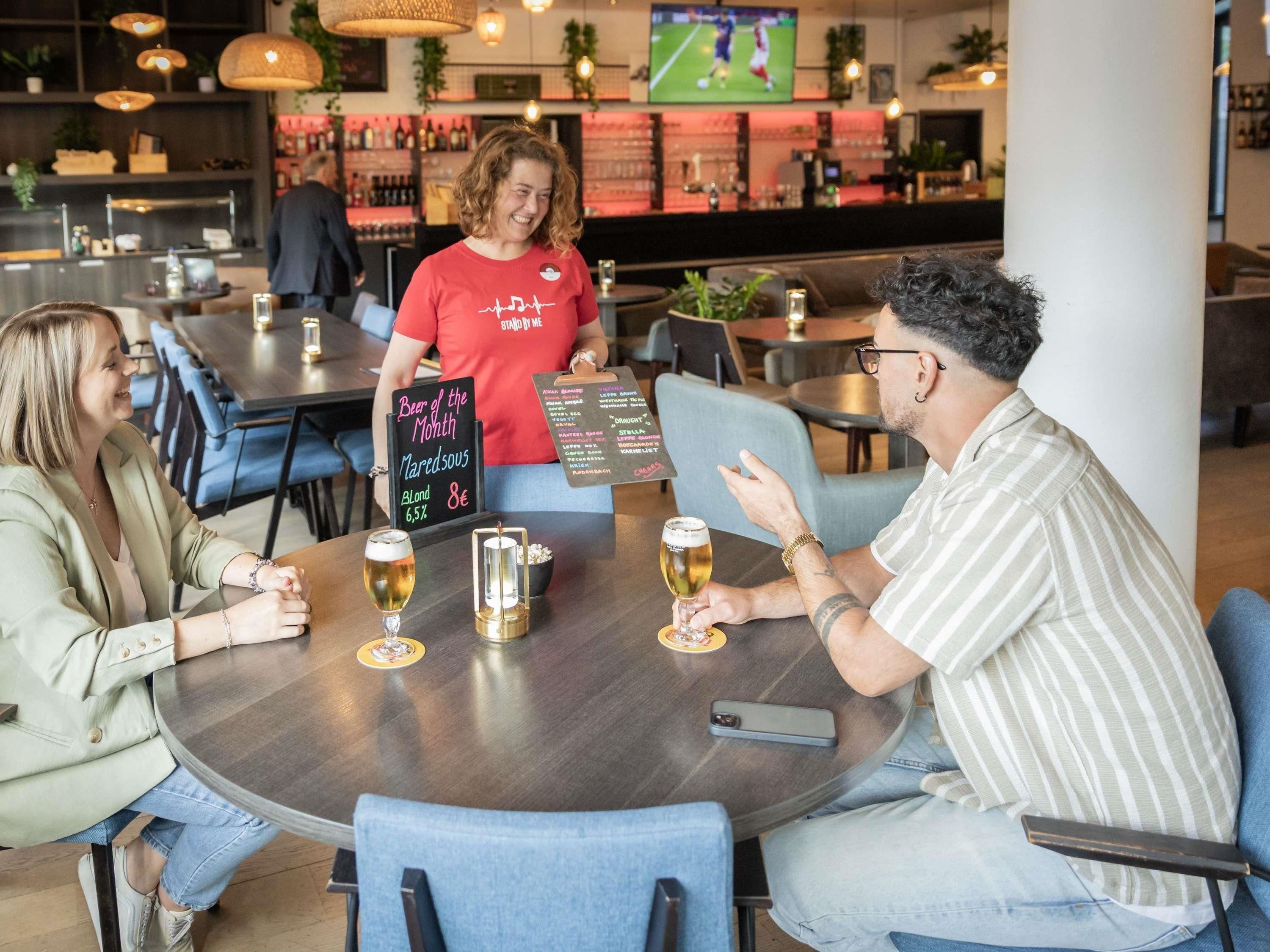 A group of people seated around a round table in a restaurant with a menu board and a chalkboard sign indicating a beer of the month.