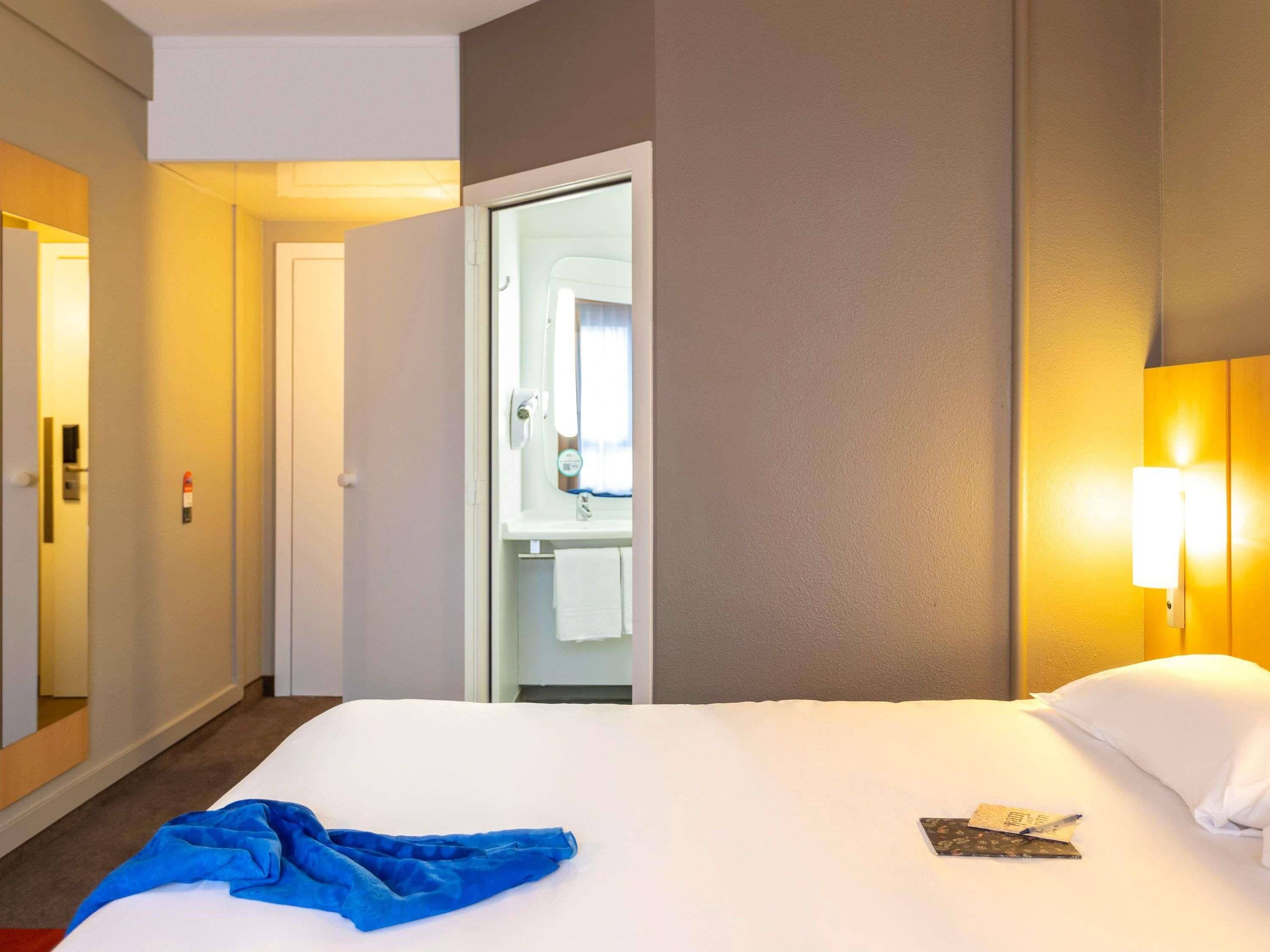 A hotel room with a bed, a bathroom visible through an open door, and a wall-mounted light fixture.