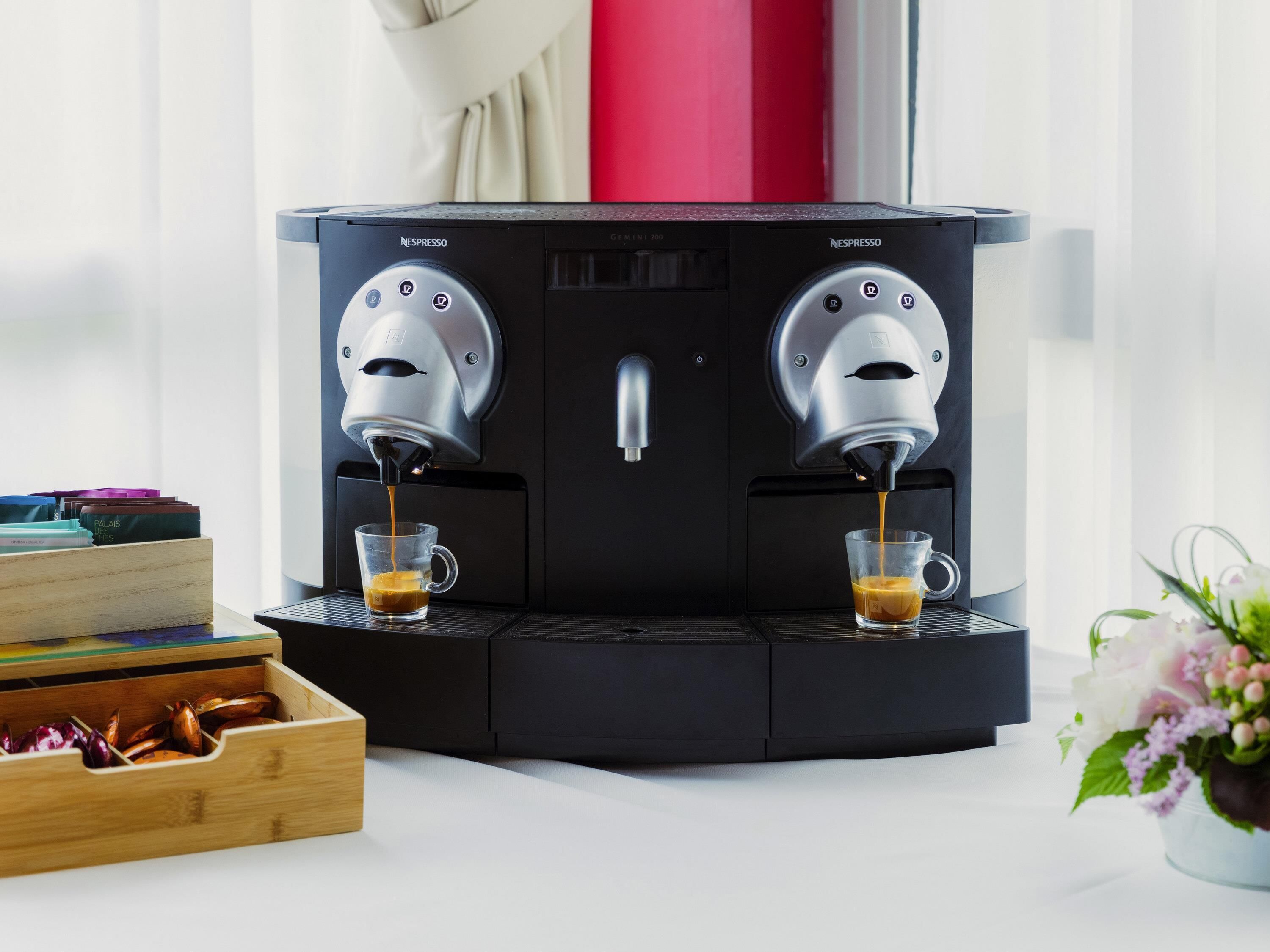 En Nespresso-kaffemaskine med to dispensere, en trækasse med kaffekapsler og en vase med blomster.