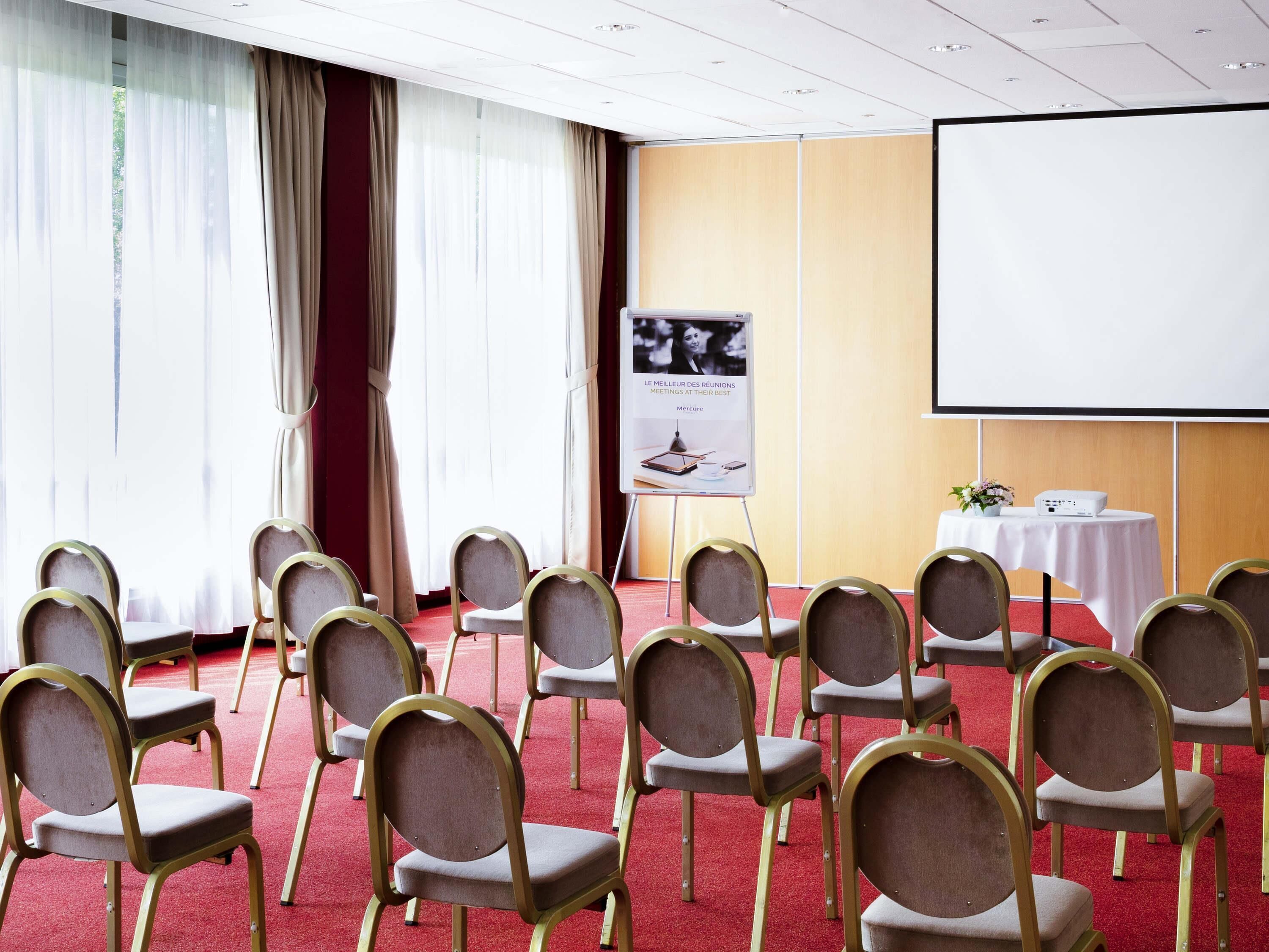 Een vergaderzaal met rijen stoelen, een projectiescherm, een whiteboard en een tafel met microfoon en bloemen.