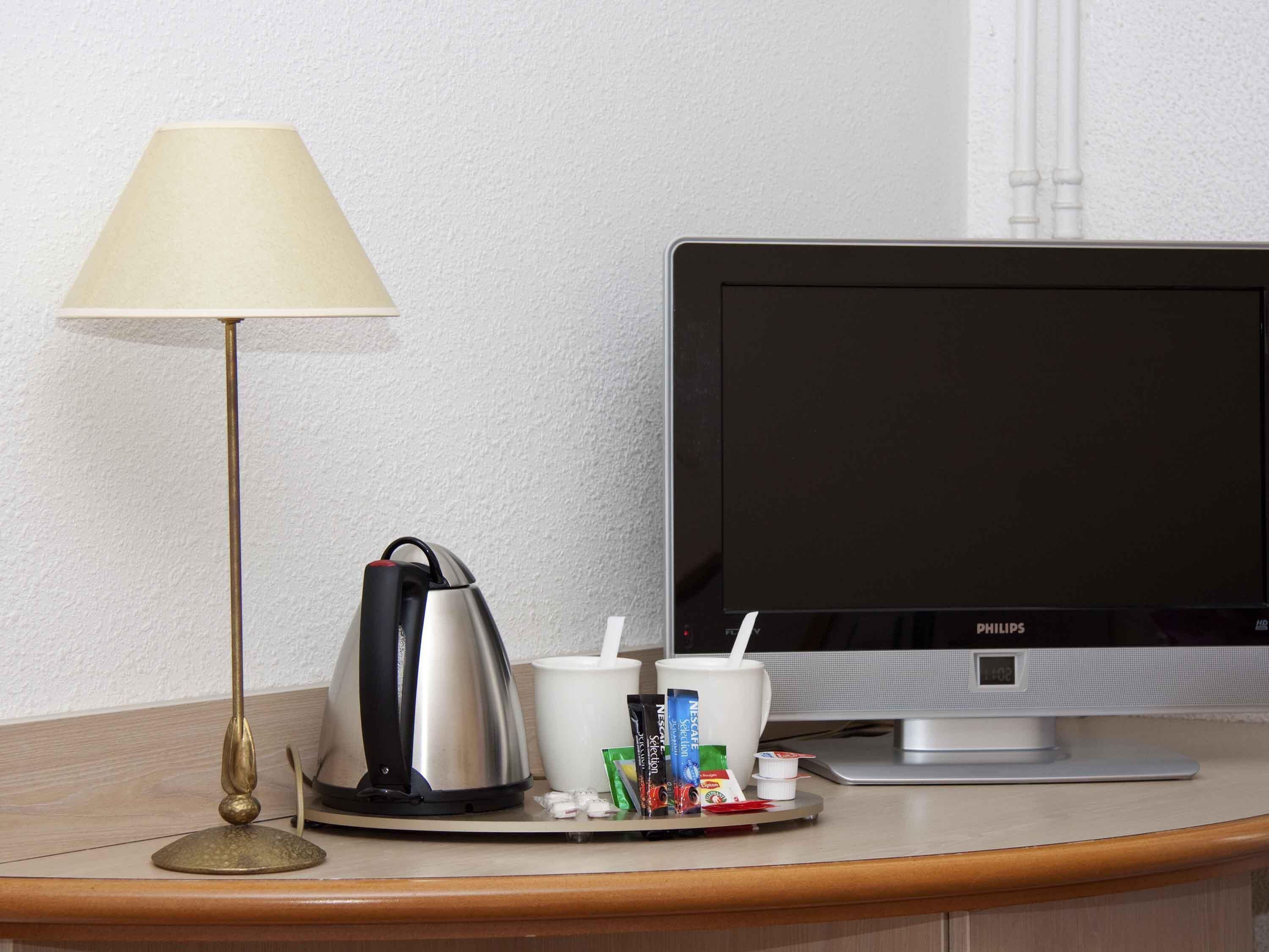 A hotel room with a desk, a lamp, a coffee maker, a TV, and a tray with condiments.