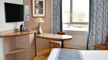 Quarto de hotel com uma cama, uma mesa com cadeira, televisão, abajur e janela com cortinas.