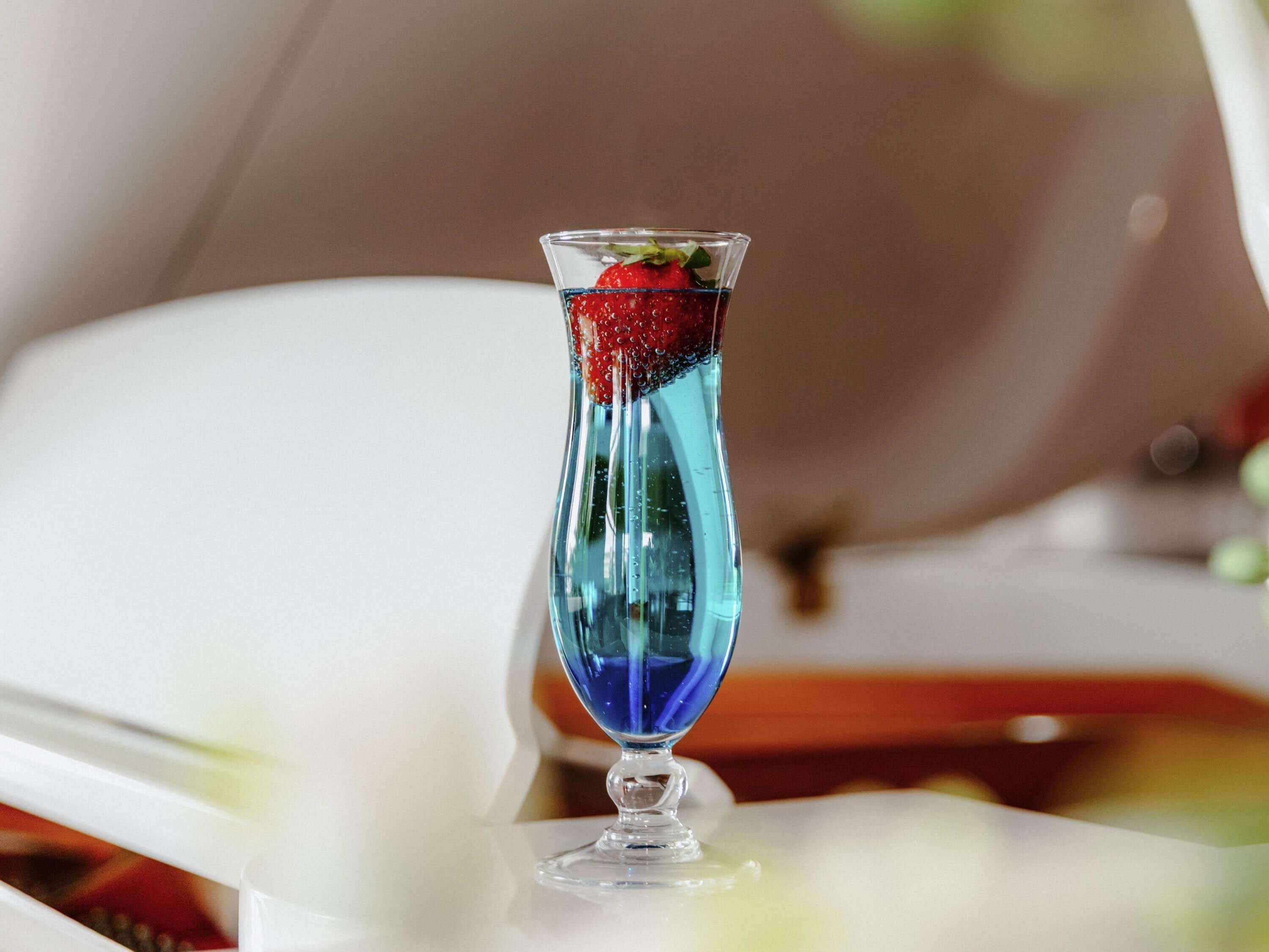 A clear glass with a blue liquid and a strawberry garnish on a white surface.