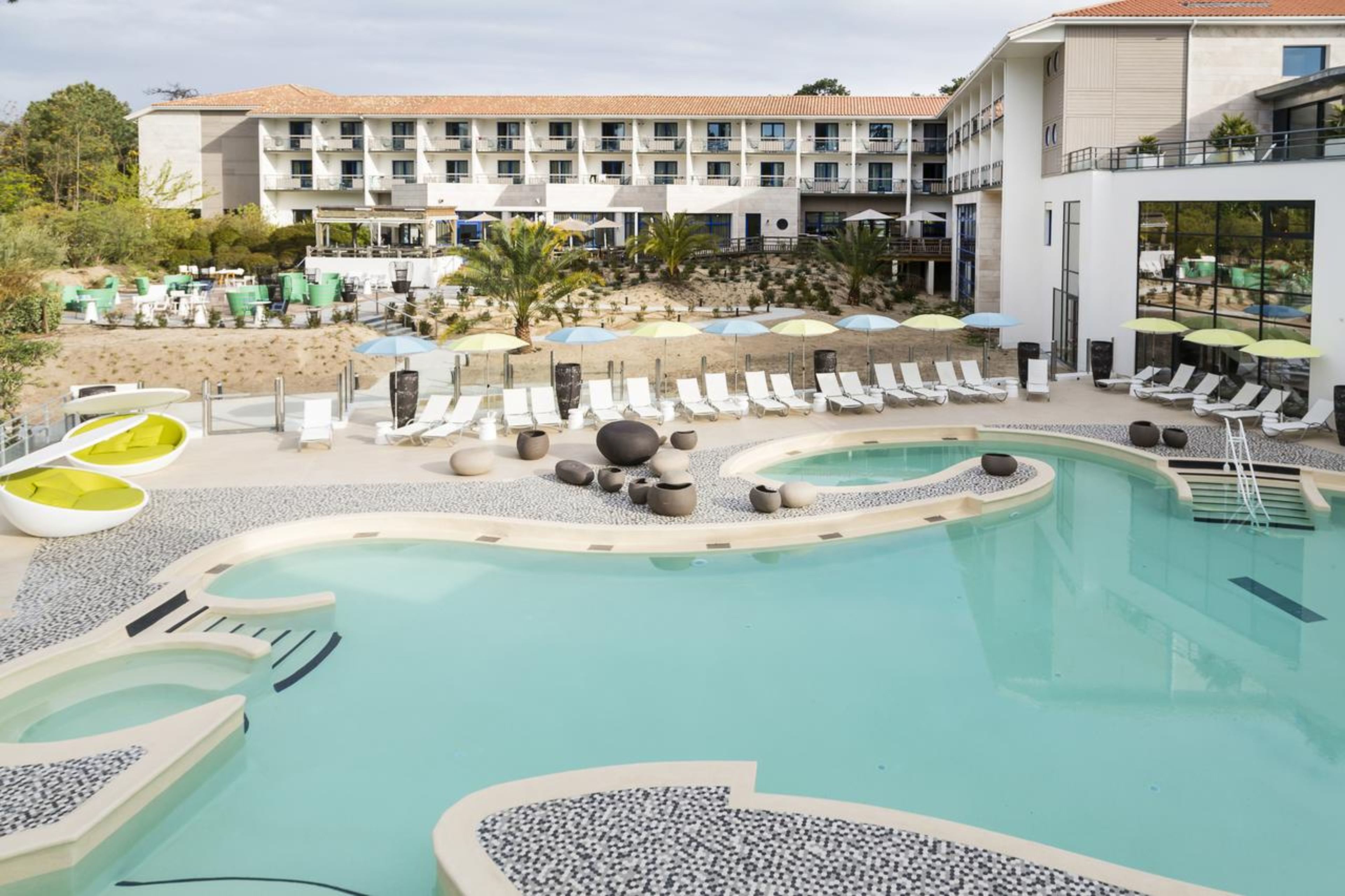 A pool area with lounge chairs and umbrellas, a building with multiple balconies, and a beach in the background.