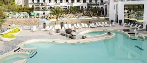 A pool area with lounge chairs and umbrellas, a building with multiple balconies, and a beach in the background.