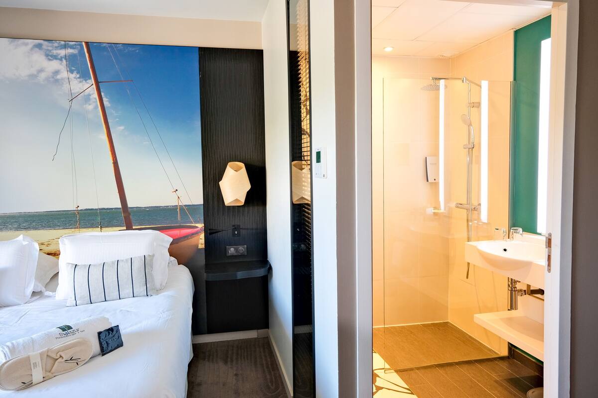Ein Hotelzimmer mit Bett, einer kleinen Sitzecke, Blick aufs Meer und einem Badezimmer mit Waschbecken und Spiegel.
