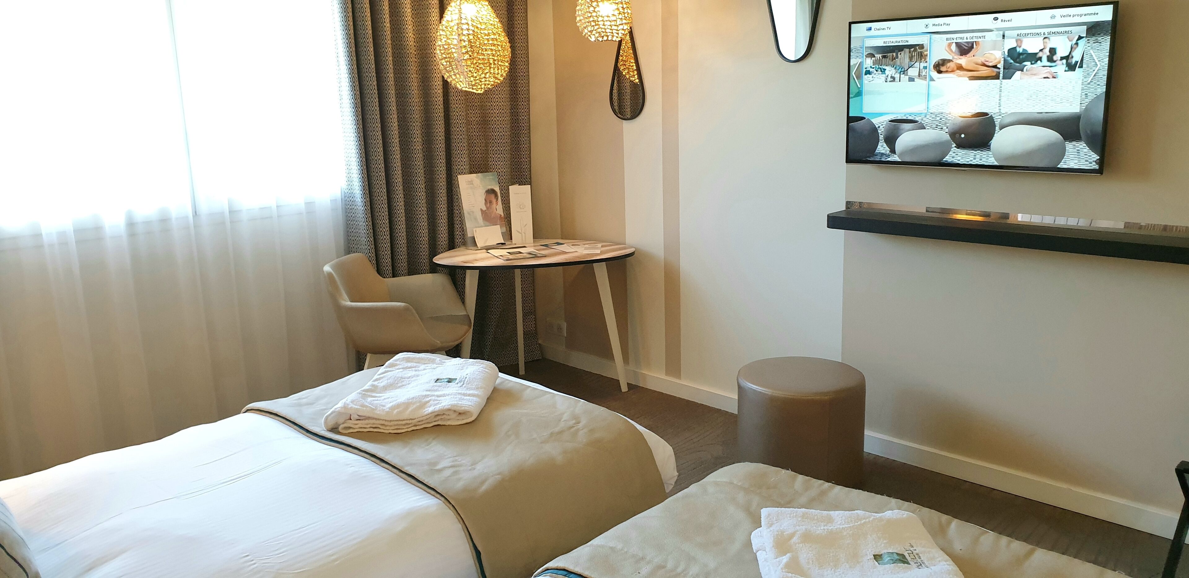 Habitación de hotel con dos camas, una mesita, una silla, televisor montado en la pared y un amplio ventanal con cortinas.