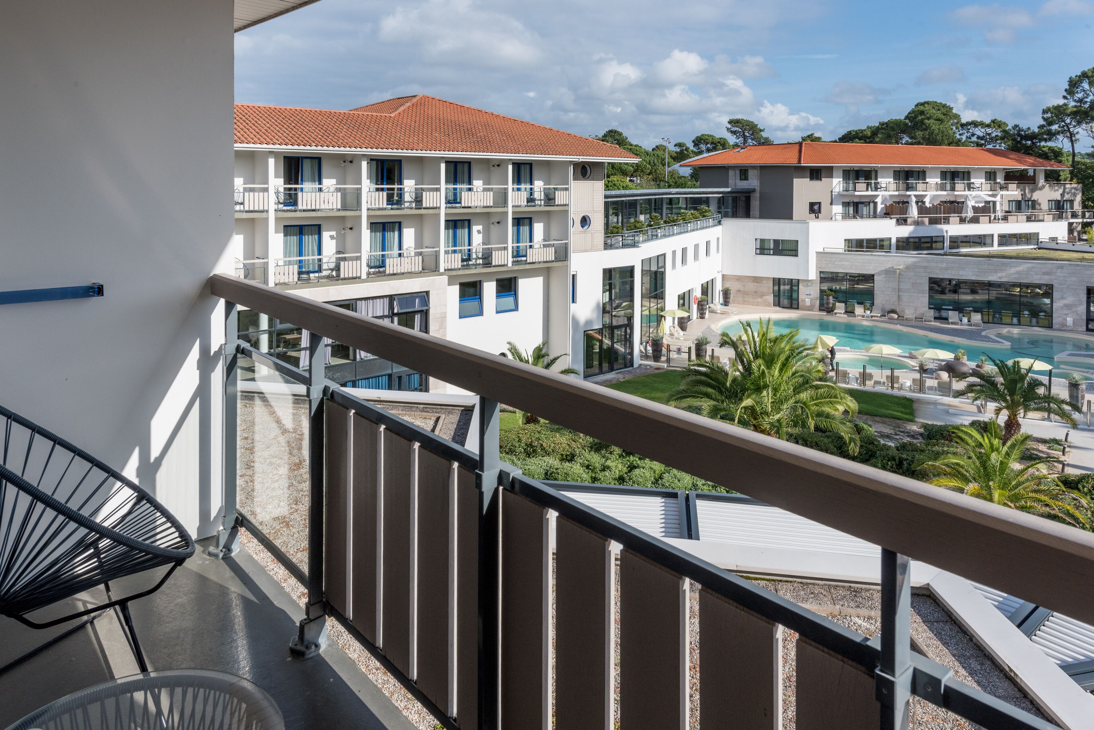 A balcony with a view of a hotel building, a swimming pool, and palm trees.
