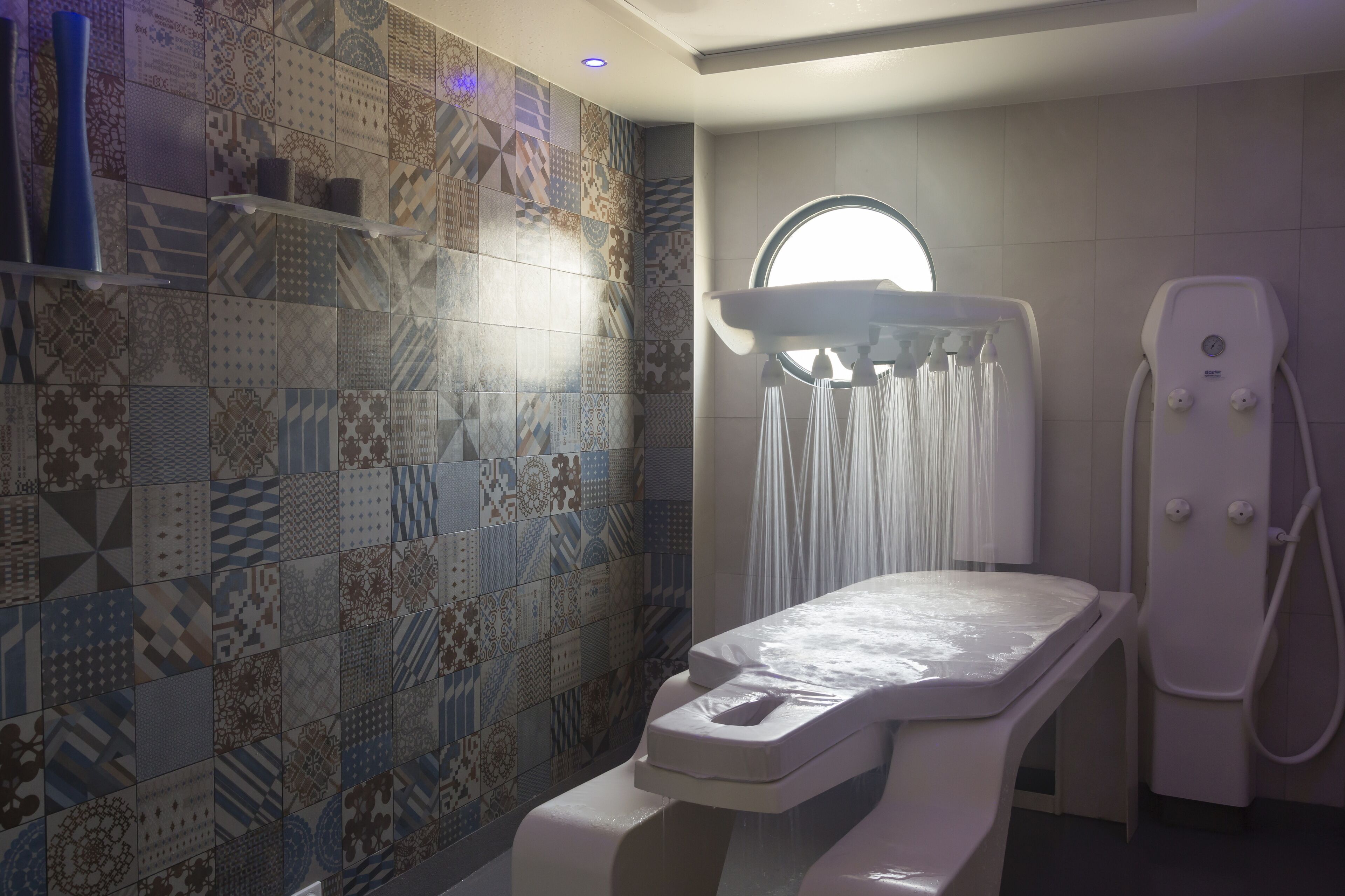 A modern bathroom with a mosaic tile wall, a standing shower, and a white examination table.