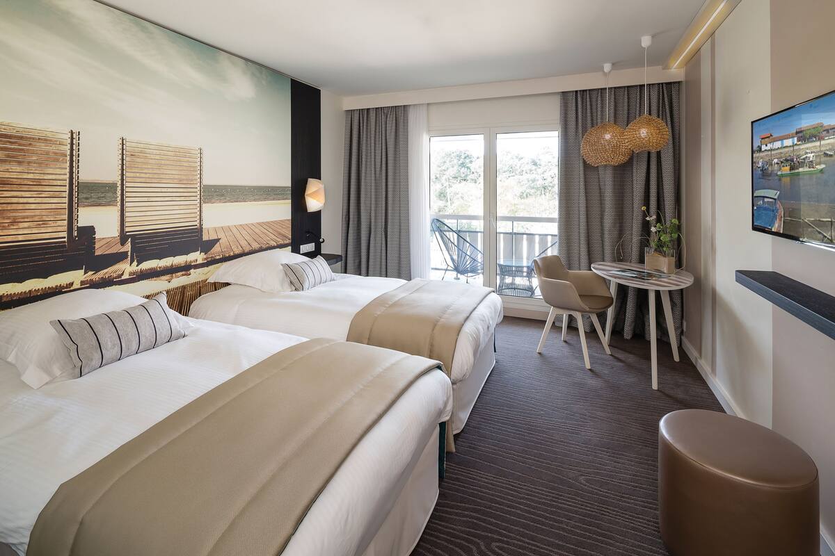 Ein Hotelzimmer mit zwei Betten, einem Balkon mit Meerblick, einem runden Tisch mit Stühlen und einem an der Wand befestigten Fernseher.