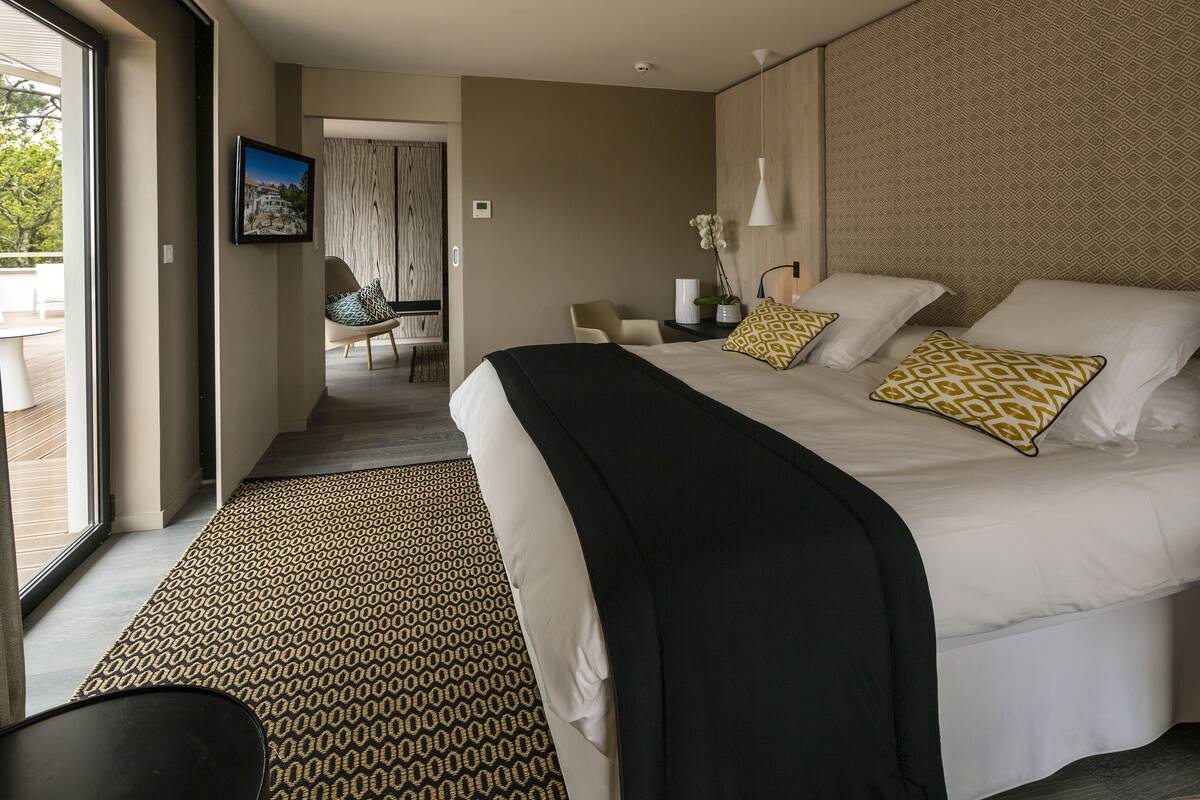 Ein modernes Hotelzimmer mit einem großen Bett, einer schwarzen Tagesdecke und gemusterten Teppichen. Es gibt einen Sessel und einen kleinen Tisch mit einer Blumenvase.