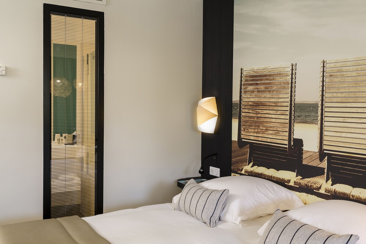 A hotel room with a bed, a nightstand, a lamp, and a view of the beach through the window.