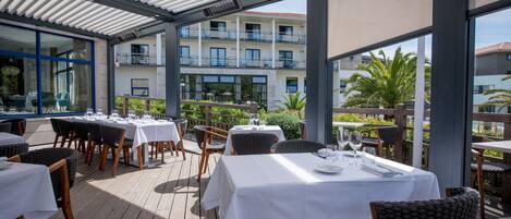 Un espace extérieur aménagé pour les repas, avec des tables et des chaises, une terrasse couverte et une vue sur un immeuble doté de balcons.