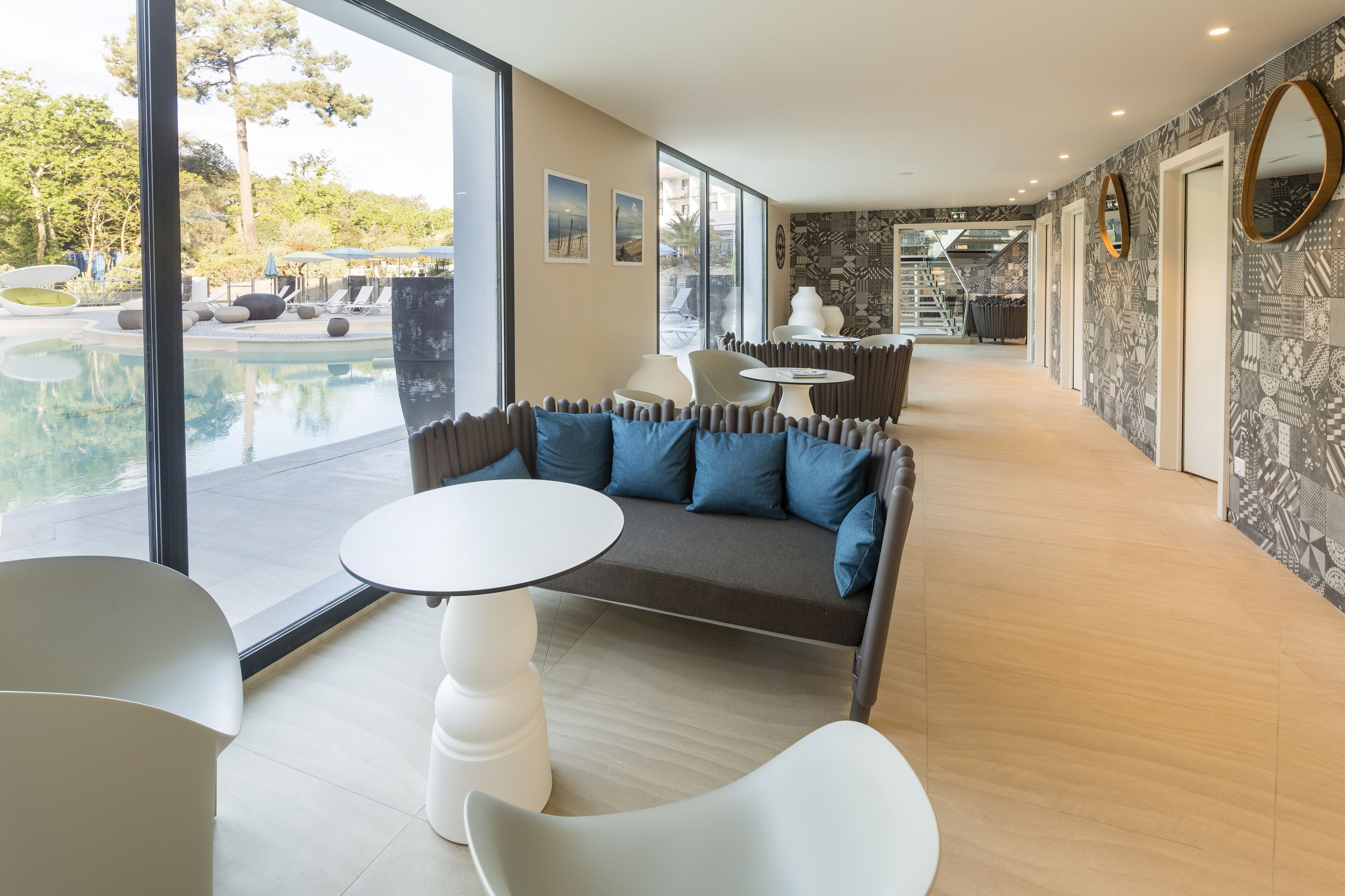 A modern hotel lobby with a pool view, a sofa with blue cushions, and a dining area with white chairs and tables.