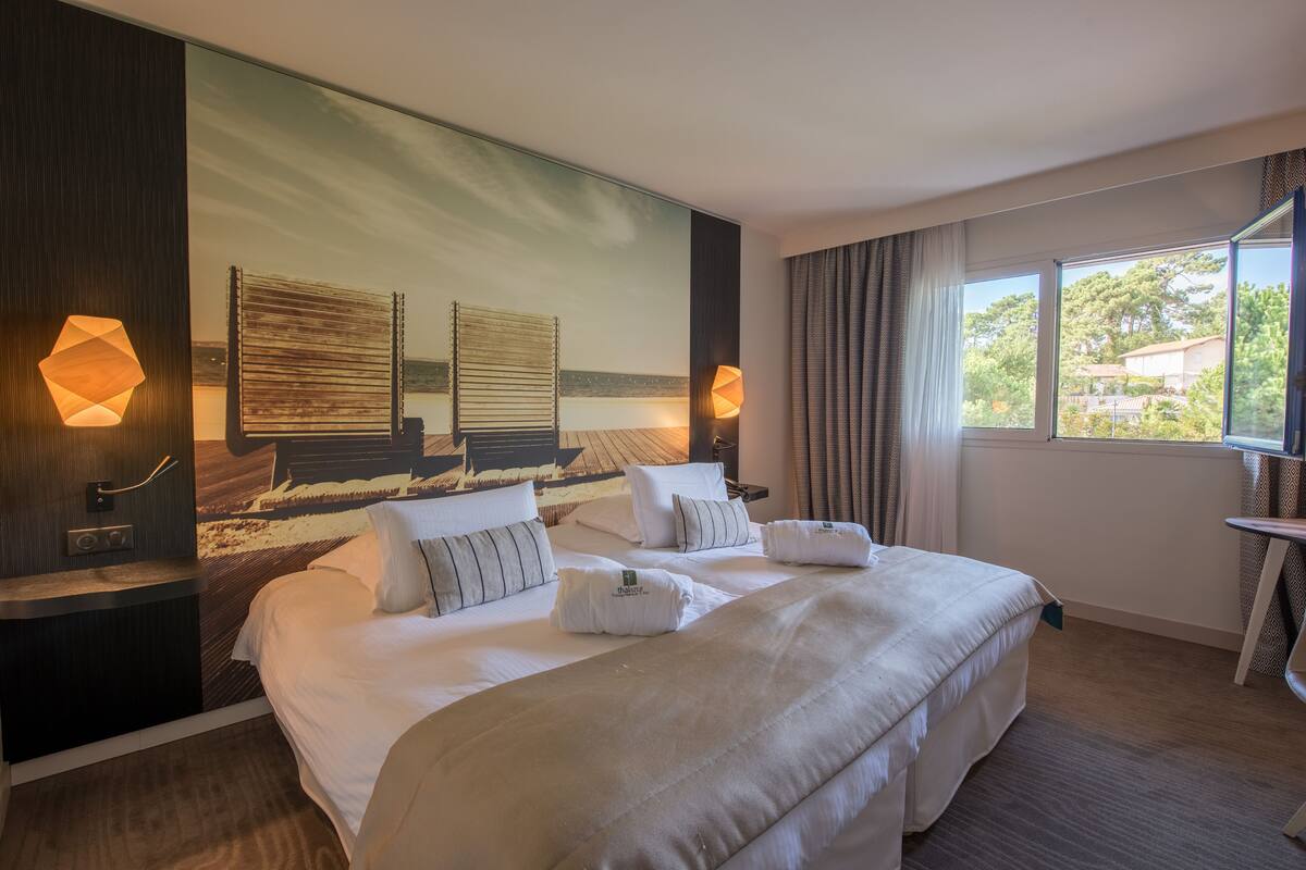 Ein Hotelzimmer mit einem großen Bett, zwei Nachttischen, einem an der Wand befestigten Fernseher und einem Fenster mit Vorhängen.