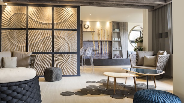 A modern living room with a sectional sofa, a coffee table, and a seating area with a round table. There is a decorative wall with a geometric pattern and a bookshelf with decorative items. - Hôtel & Spa - Thalazur Arcachon (Arcachon)