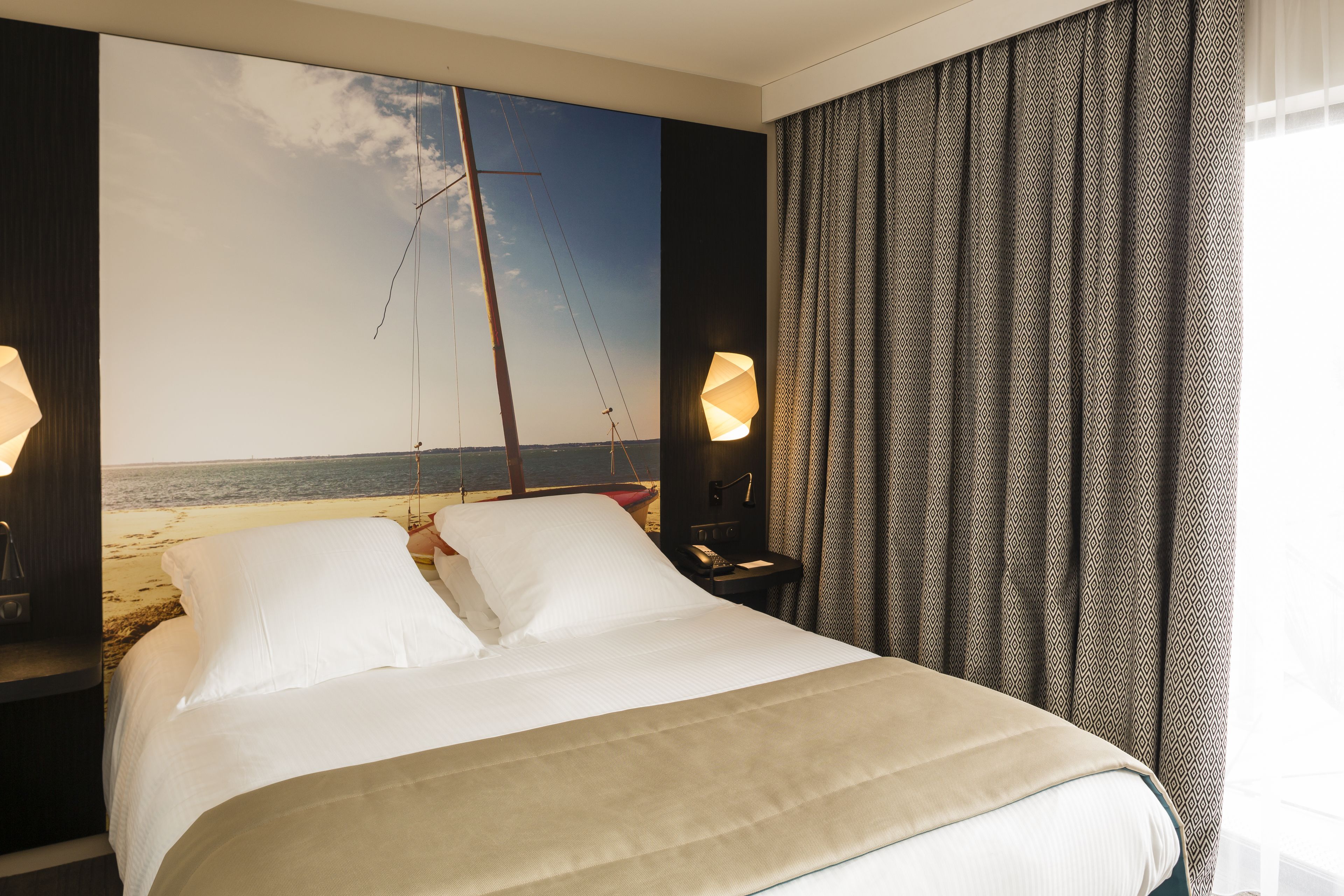 A hotel room with a bed, a nightstand, a lamp, and a view of the beach.